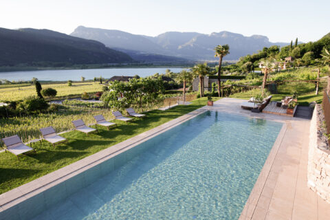 Infinity-Pool am Hang mit Liegen und Rasen, Blick über den Kalterer See zu Bergen unter klarem Himmel