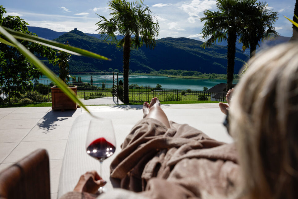 Person mit Bademantel und ausgestreckten Beinen auf Terrasse, hält Weinglas; Palmen, See und Berge im Hintergrund.