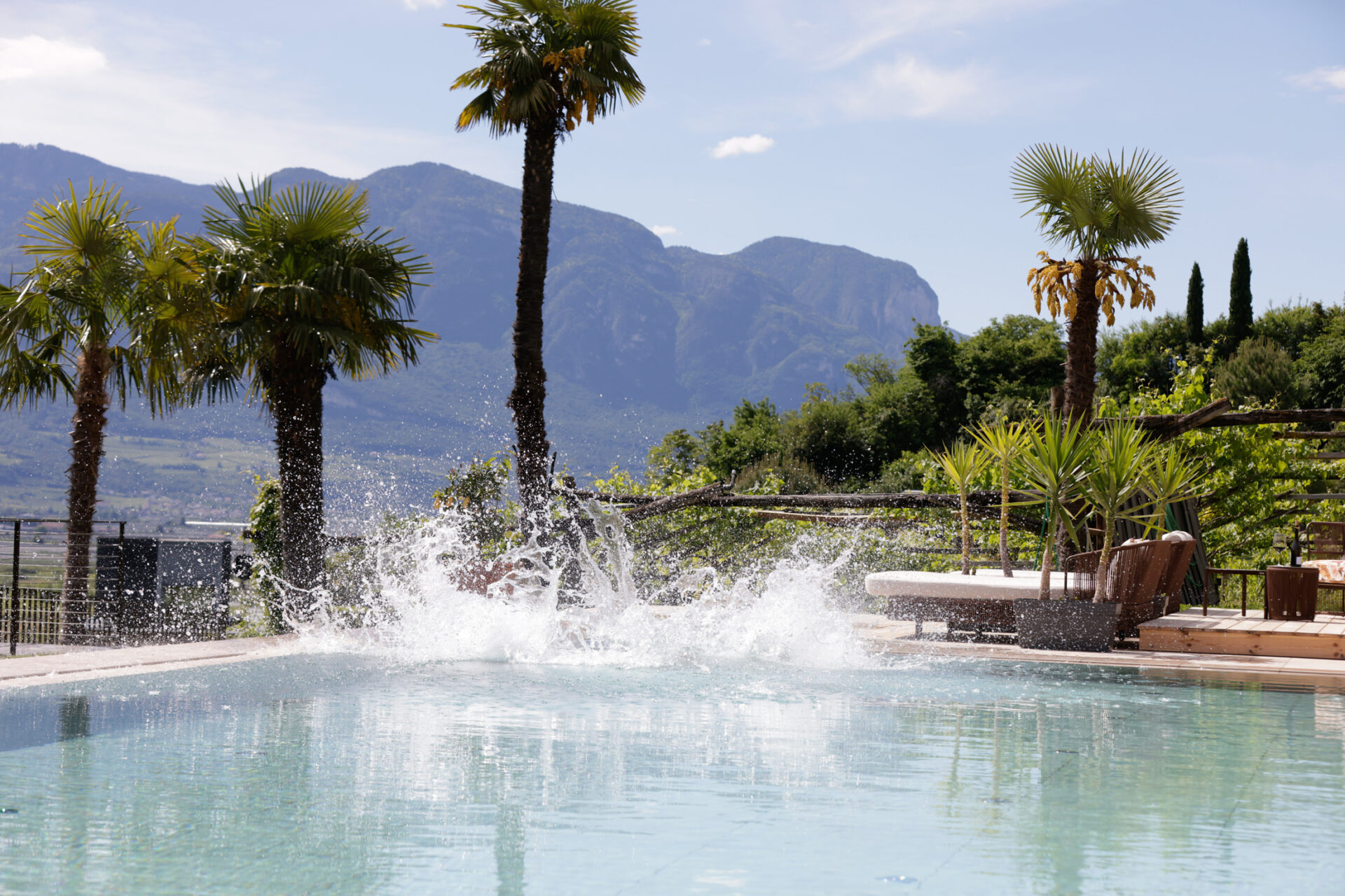 Spritzender Wassereffekt in freiem Pool mit Palmen, Liegebereich und Bergen im Hintergrund, Hotelpool am Kalterer See.