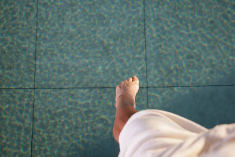 Nackter Fuß und Unterschenkel hängen über türkis gemusterten Poolfliesen; weißes Tuch am Oberschenkel.