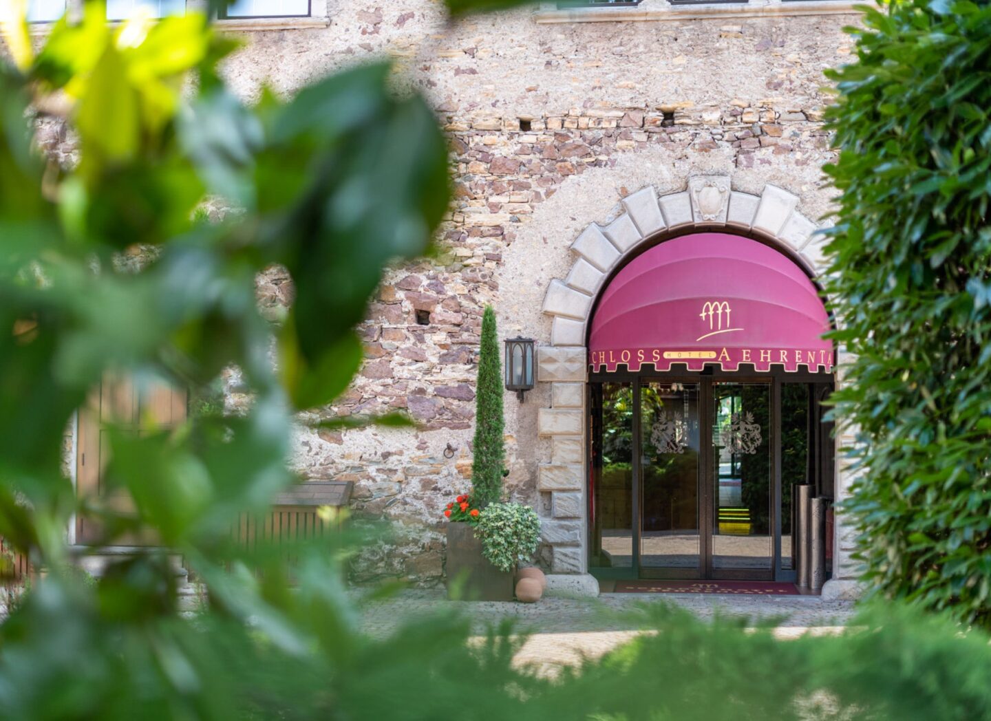 Hotelfassade: Steinmauer mit rundem Torbogen und magentafarbenem Vordach, Wandlaternen und Pflanzen im Vordergrund