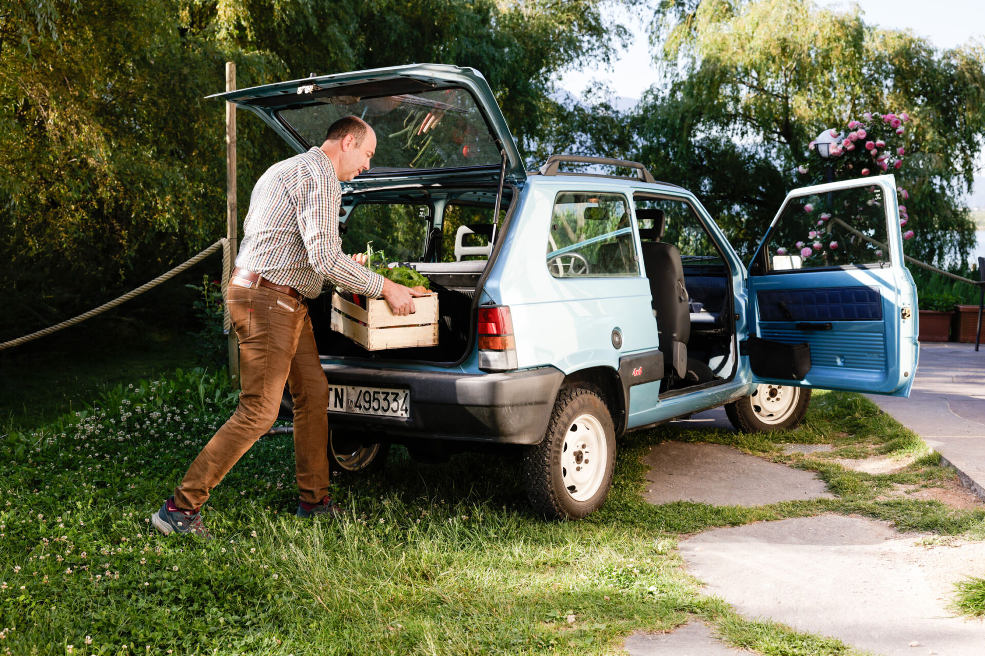 Mann lädt Kiste in Kofferraum eines kleinen blauen Autos; zweite Person an geöffneter Tür; grasiger Platz mit Bäumen.