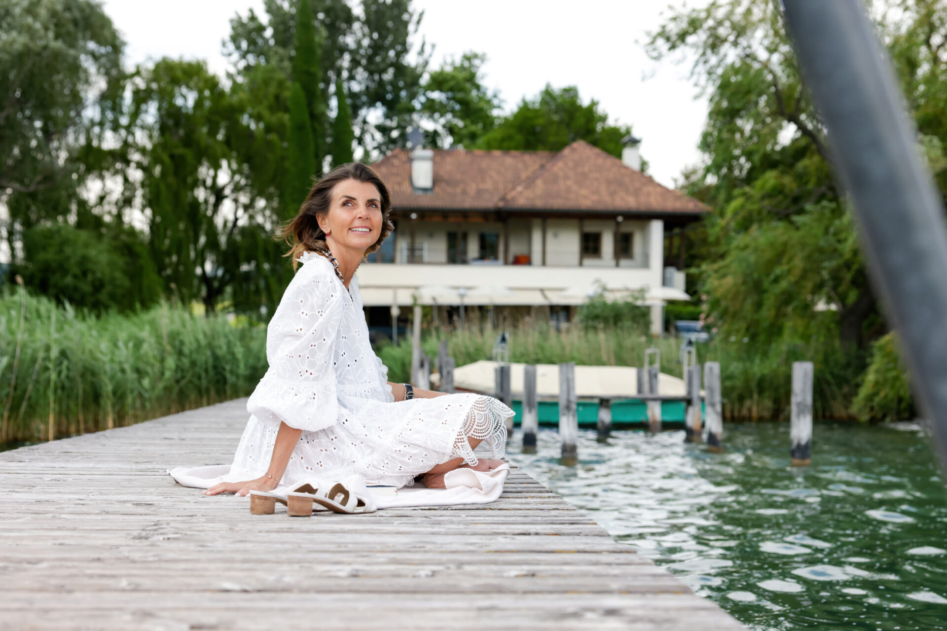 Frau in weißem Kleid sitzt barfuß auf einem Holzsteg am See, lächelnd; im Hintergrund ein zweistöckiges Haus, Bojen und Uferv