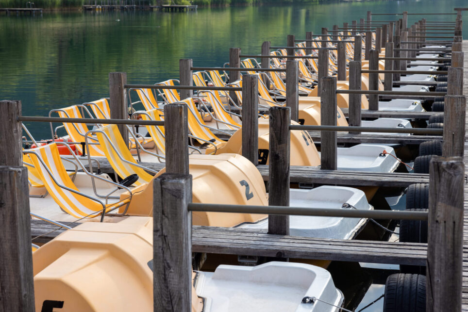 Mehrere Tretboote mit gelben Sonnendächern an einem Holzsteg, ruhiges Wasser und Ufer mit Bäumen (Kalterer See)
