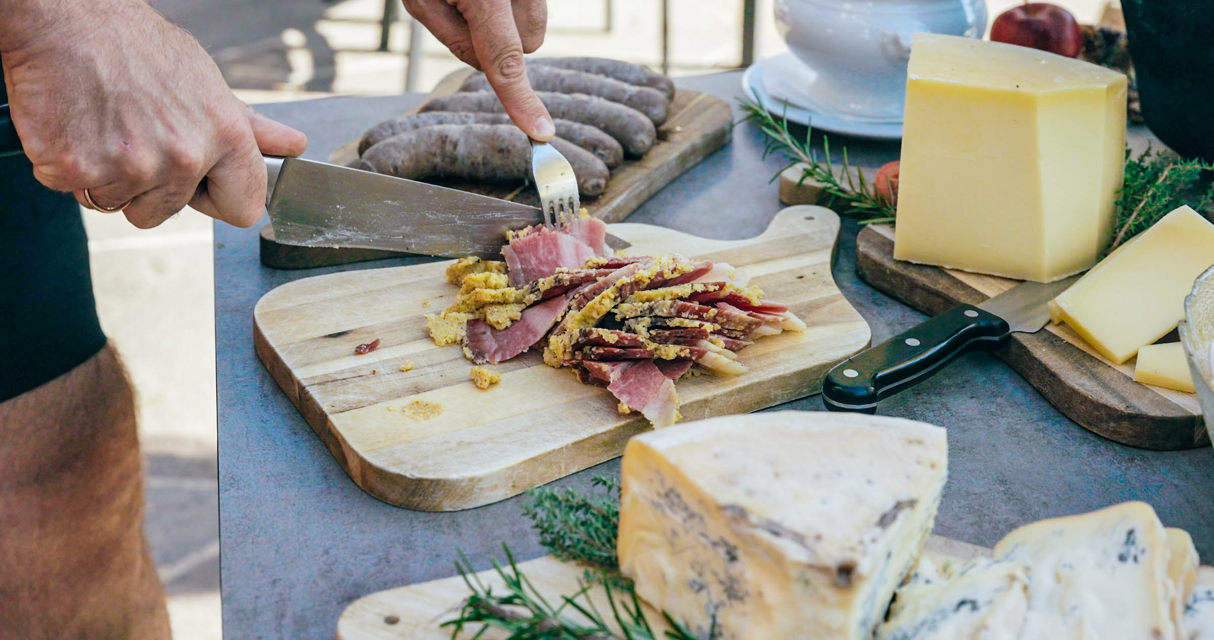 Schneidebrett mit geschnittenem gepökeltem Fleisch, Käsestück, Würstchen im Hintergrund und Kräutern; Hand schneidet