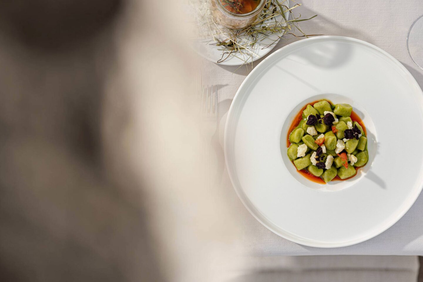 Runder weißer Teller mit klein gewürfeltem Gemüse in heller Sauce, auf hellem Tisch; oben Glas und Stoffserviette