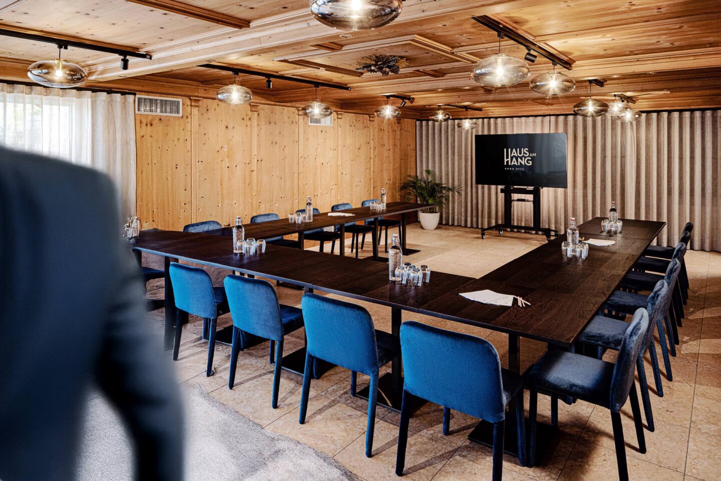 Tagungsraum mit Holzverkleidung, U-förmiger Besprechungstisch, blaue Stühle, Monitor und Deckenlampen.