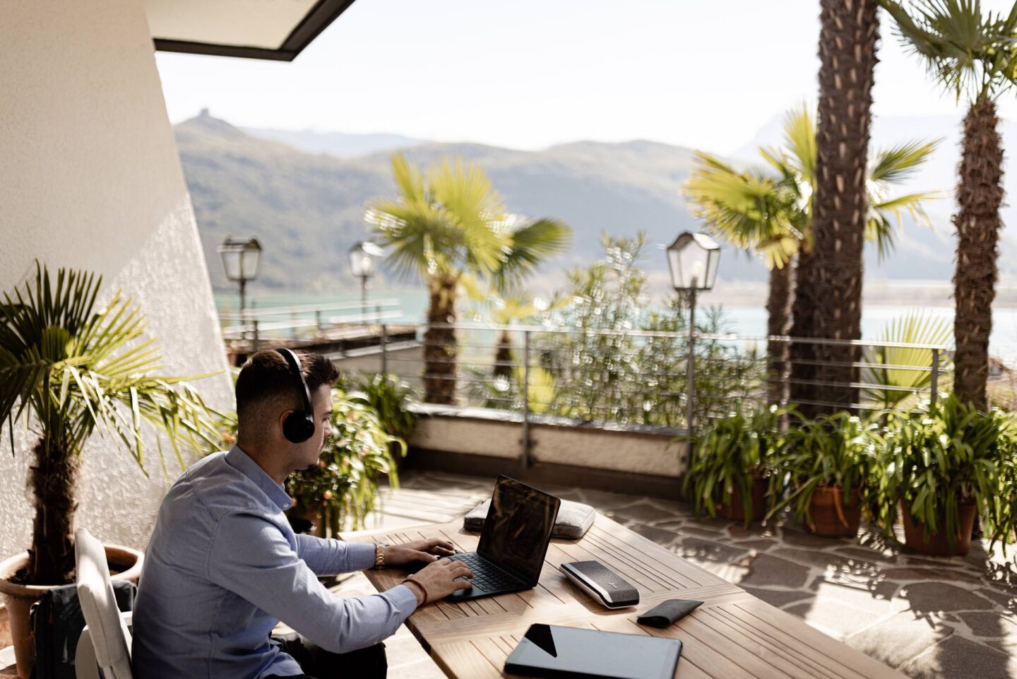Person mit Kopfhörern am Laptop auf Terrasse eines Tagungshotels, Palmen, Blick auf Kalterer See und Berge
