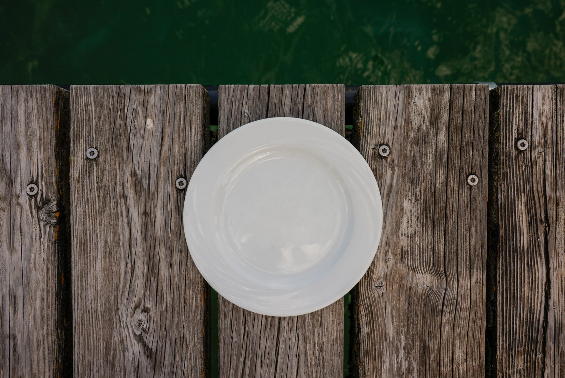 Weißer Teller auf verwittertem Holzsteg, grünes Seewasser im Hintergrund