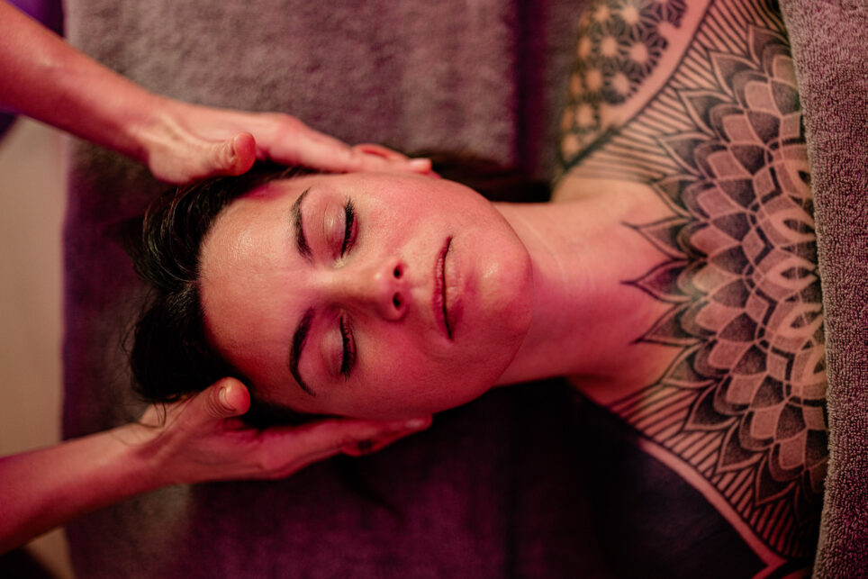 Entspannte Person mit geschlossenen Augen erhält Kopfmassage, sichtbare geometrische Brusttattoos, warmes rotes Licht.