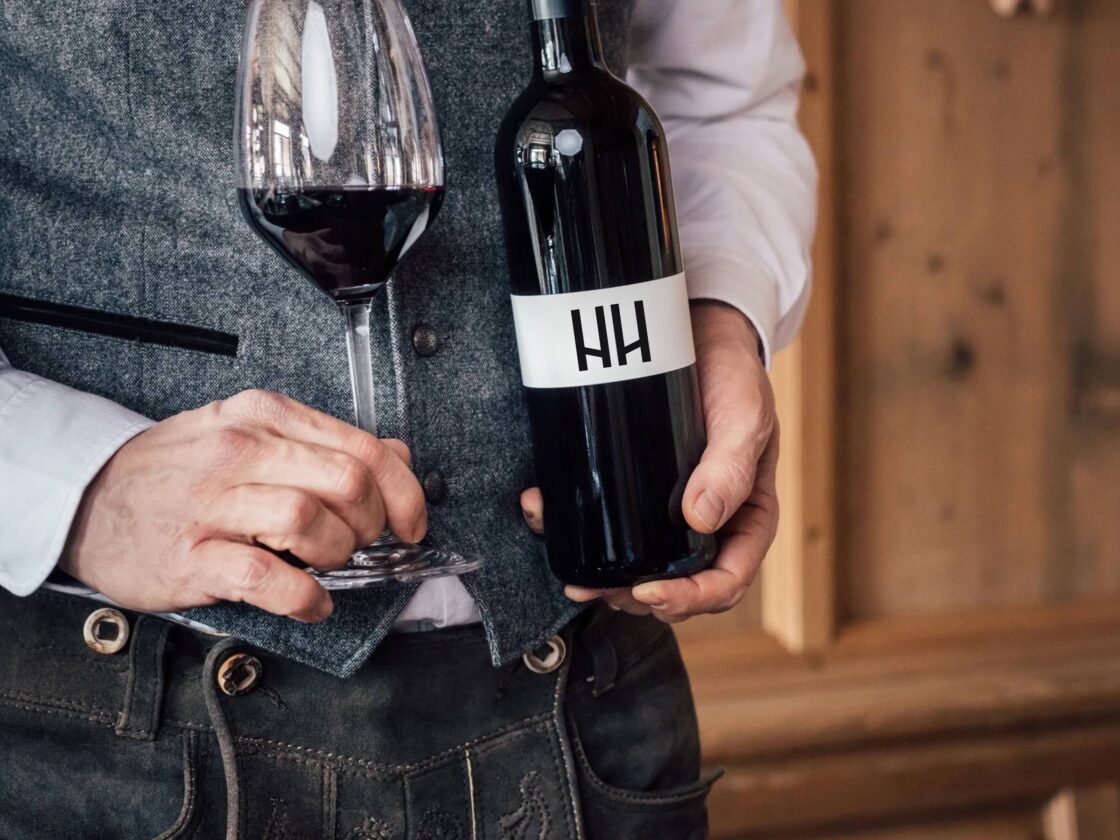 Hände einer Person halten eine geöffnete Rotweinflasche mit HH-Etikett und ein halbvolles Glas vor Holzverkleidung.