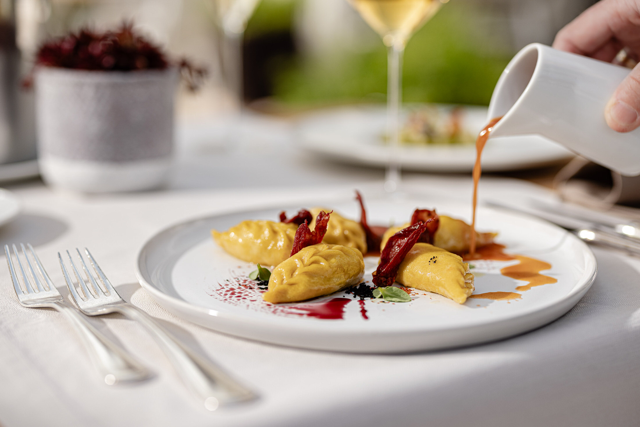 Gefüllte Pasta auf weißem Teller; braune Sauce aus Kännchen, rote Beilage und Kräuter; Besteck und Weingläser unscharf