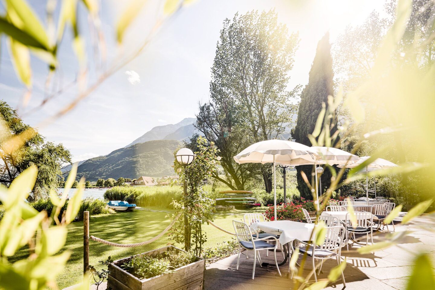 Terrasse mit Tischen, Sonnenschirmen und Stühlen auf einer Wiese, dahinter hohe Bäume und bergige Landschaft im Gegenlicht.