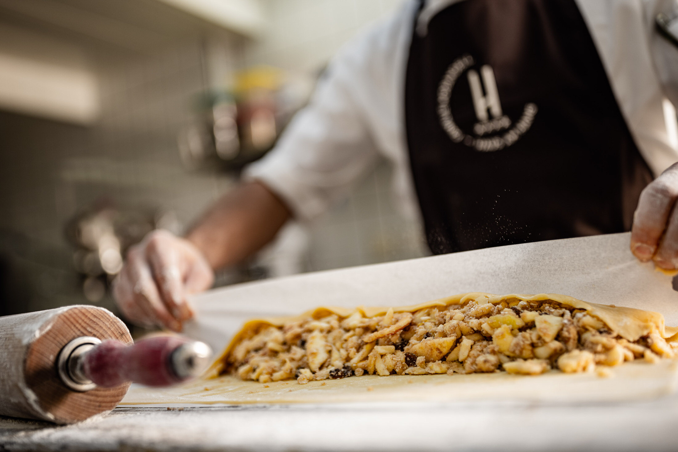 Hände rollen gefüllten Teigstrang auf Tisch in Restaurantküche; grobe Stückchenfüllung, Person mit Schürze im Hintergrund