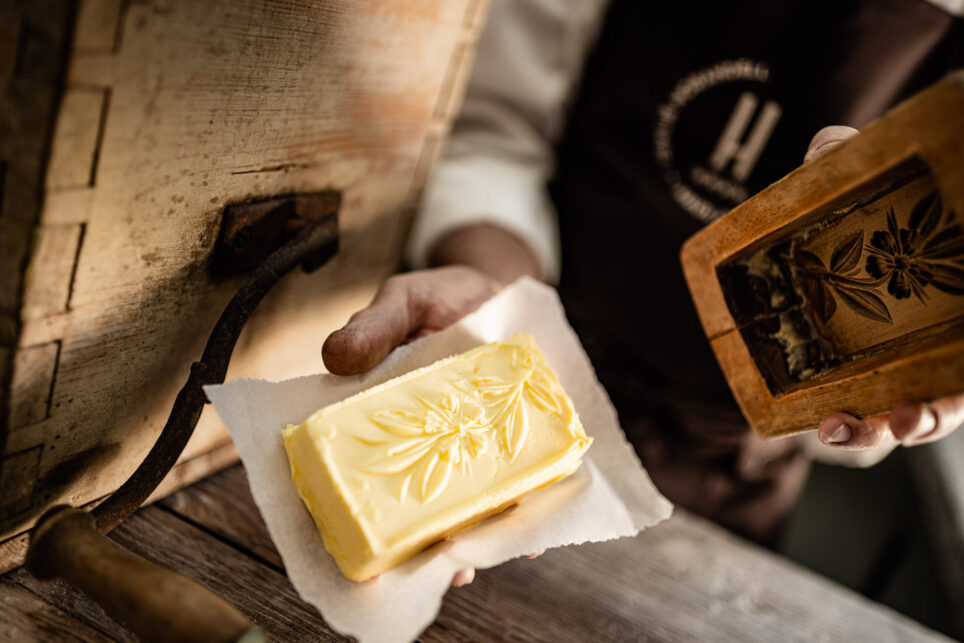 Hand in Schürze hält Butter mit Blattprägung auf Serviette; daneben Flasche auf rustikalem Holztisch