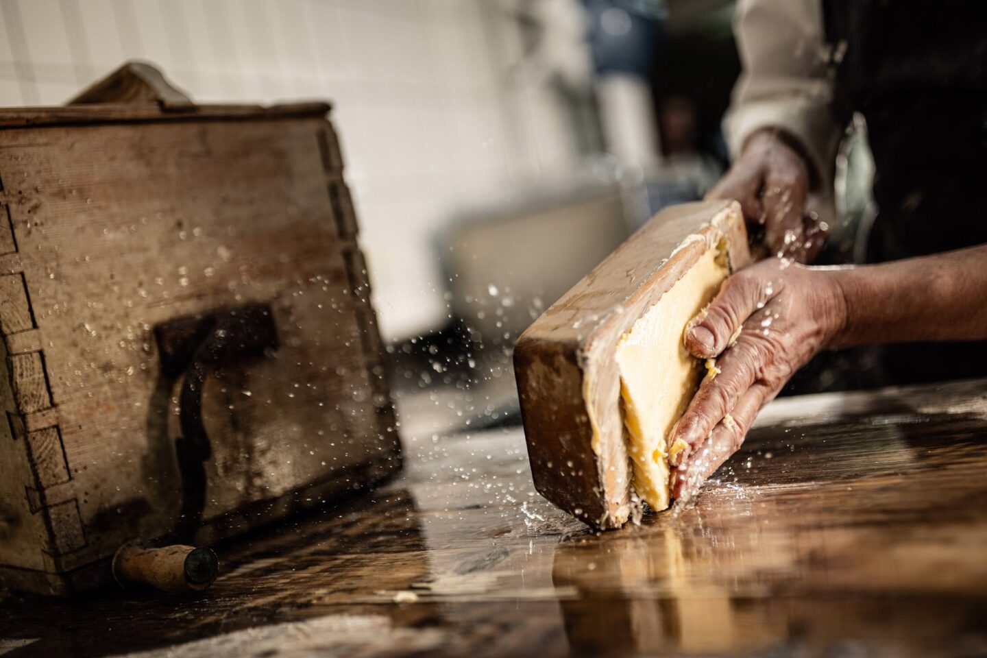 Hände schneiden ein Stück gereiften Käse vom Laib auf nassem Holztisch; Krümel und Flüssigkeit spritzen.