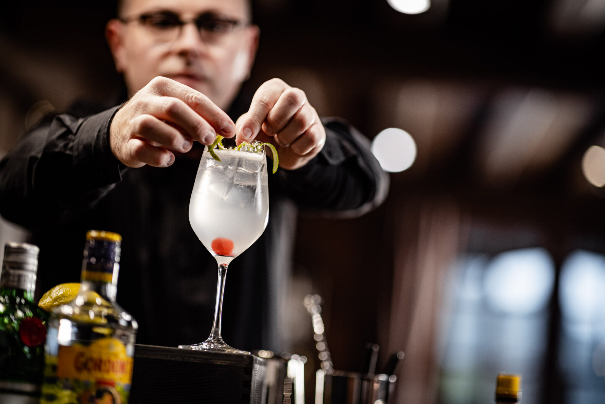 Barkeeperhände platzieren Kräutergarnitur mit Zange in Glas mit Eis und Kirsche; unscharfer Barhintergrund