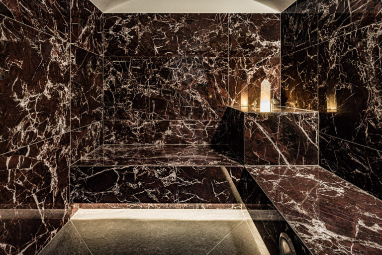 Dunkle, glänzende Marmorwände mit eingelassener Ablage, drei brennende Kerzen und indirekte Bodenbeleuchtung