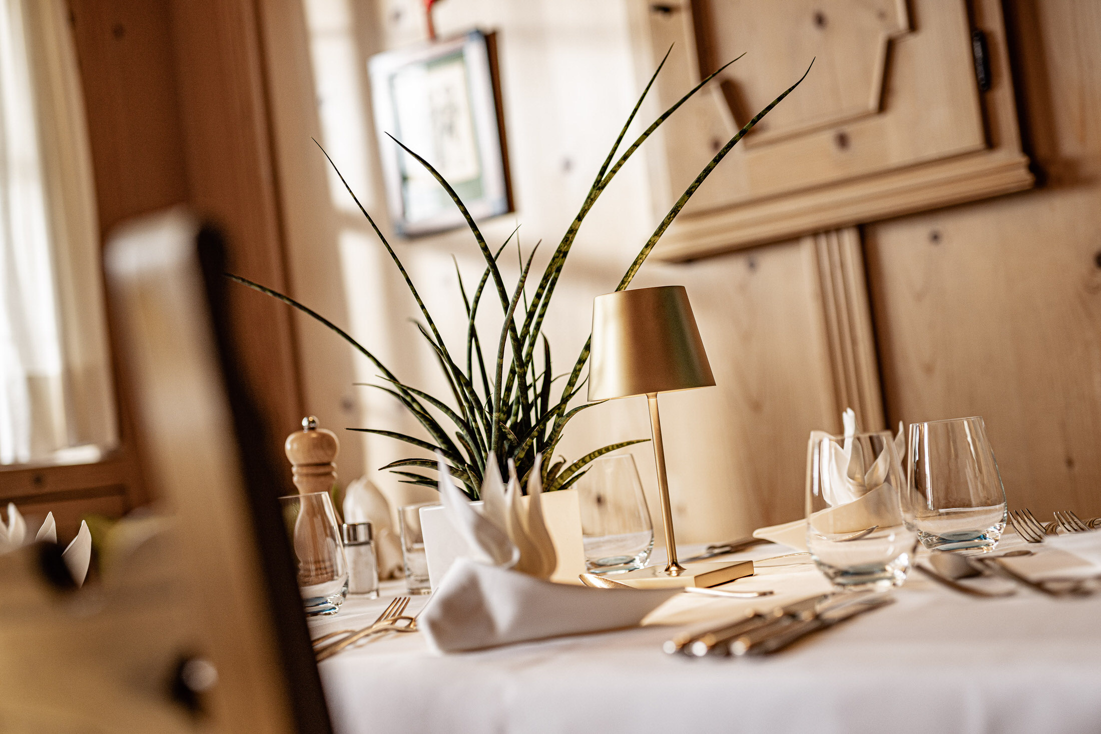 Gedeckter Tisch im Restaurant mit Holzvertäfelung, Topfpflanze, gefalteten Stoffservietten, Besteck und Tischlampe