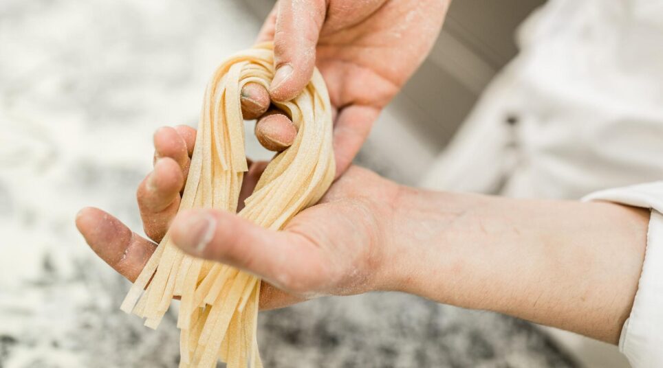 Zwei Hände halten frisch geschnittene Bandnudeln aus Teig, Hintergrund unscharf.
