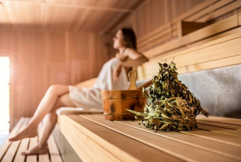 Person mit Handtuch sitzt auf Holzbank in Hotelsauna; Saunakübel und Aufgussbesen aus Birkenzweigen im Vordergrund.
