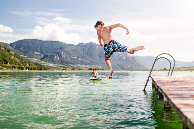 Mann springt von Holzsteg in grünes Wasser des Kalterer Sees; Berge am Horizont, weitere Person auf Steg und Leiter am Ufer.