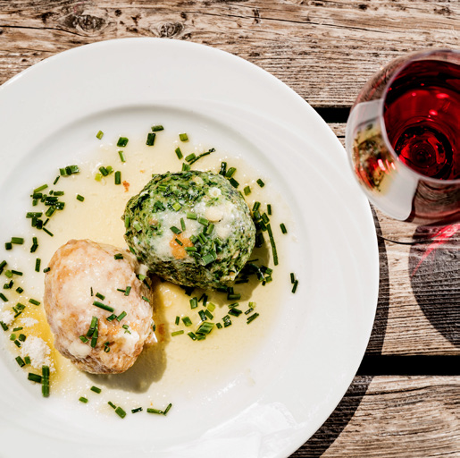 zwei runde Knödel mit Kräuterbutter und gehackter Petersilie auf weißem Teller, Holztisch, Glas Rotwein rechts